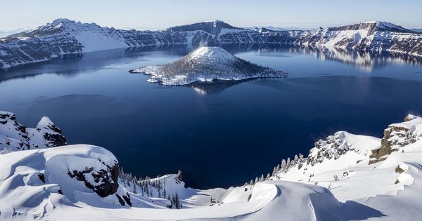 Crater Lake in winter — snow-blanketed caldera rim with the deep blue lake and Wizard Island visible below