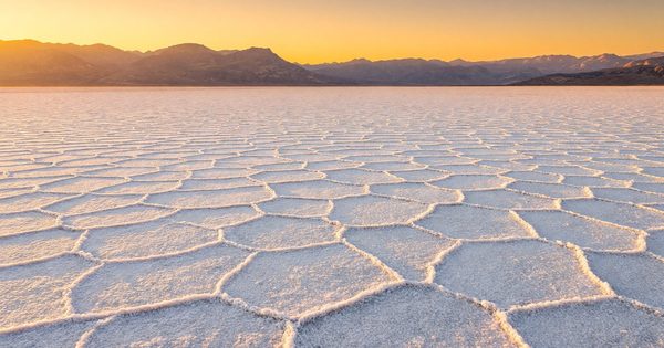 Wide panoramic view of Badwater Basin salt flats at Death Valley National Park at golden hour, crystalline white salt polygons extending to distant mountain ranges under a warm amber sky