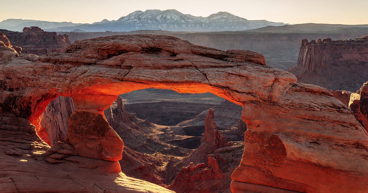 Mesa Arch at Canyonlands at sunrise — the underside of the sandstone arch glowing orange-red as first light fires through from the canyon below, with the La Sal Mountains visible in the background