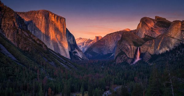 Tunnel View at Yosemite at sunset — El Capitan and Half Dome bathed in deep orange alpenglow, Bridalveil Fall catching the last warm light, Yosemite Valley floor in deep blue shadow below
