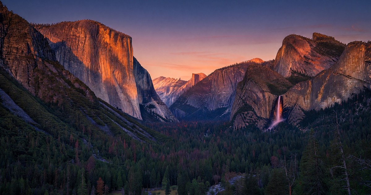 Tunnel View at Yosemite at sunset — El Capitan and Half Dome bathed in deep orange alpenglow, Bridalveil Fall catching the last warm light, Yosemite Valley floor in deep blue shadow below